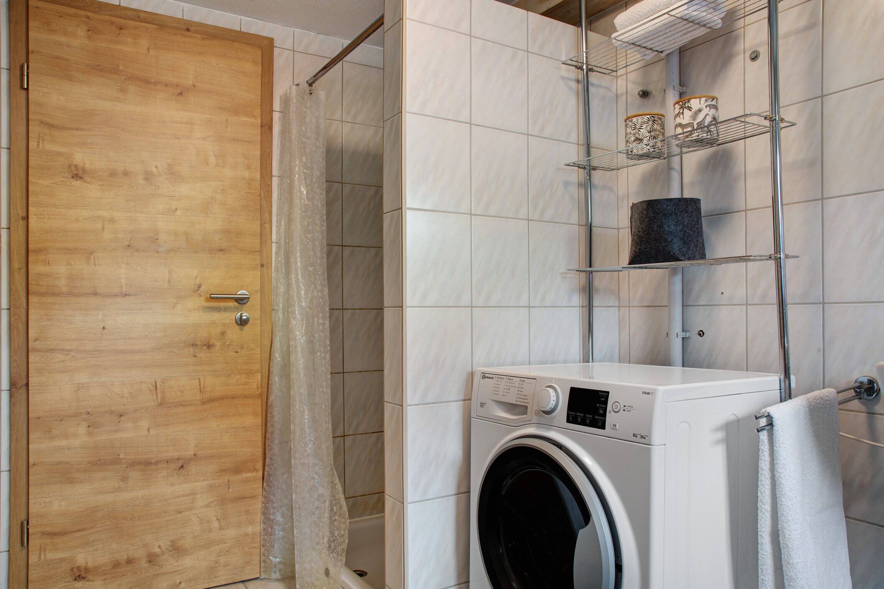 Un área de lavandería con lavadora, una puerta de madera y paredes con baldosas.
