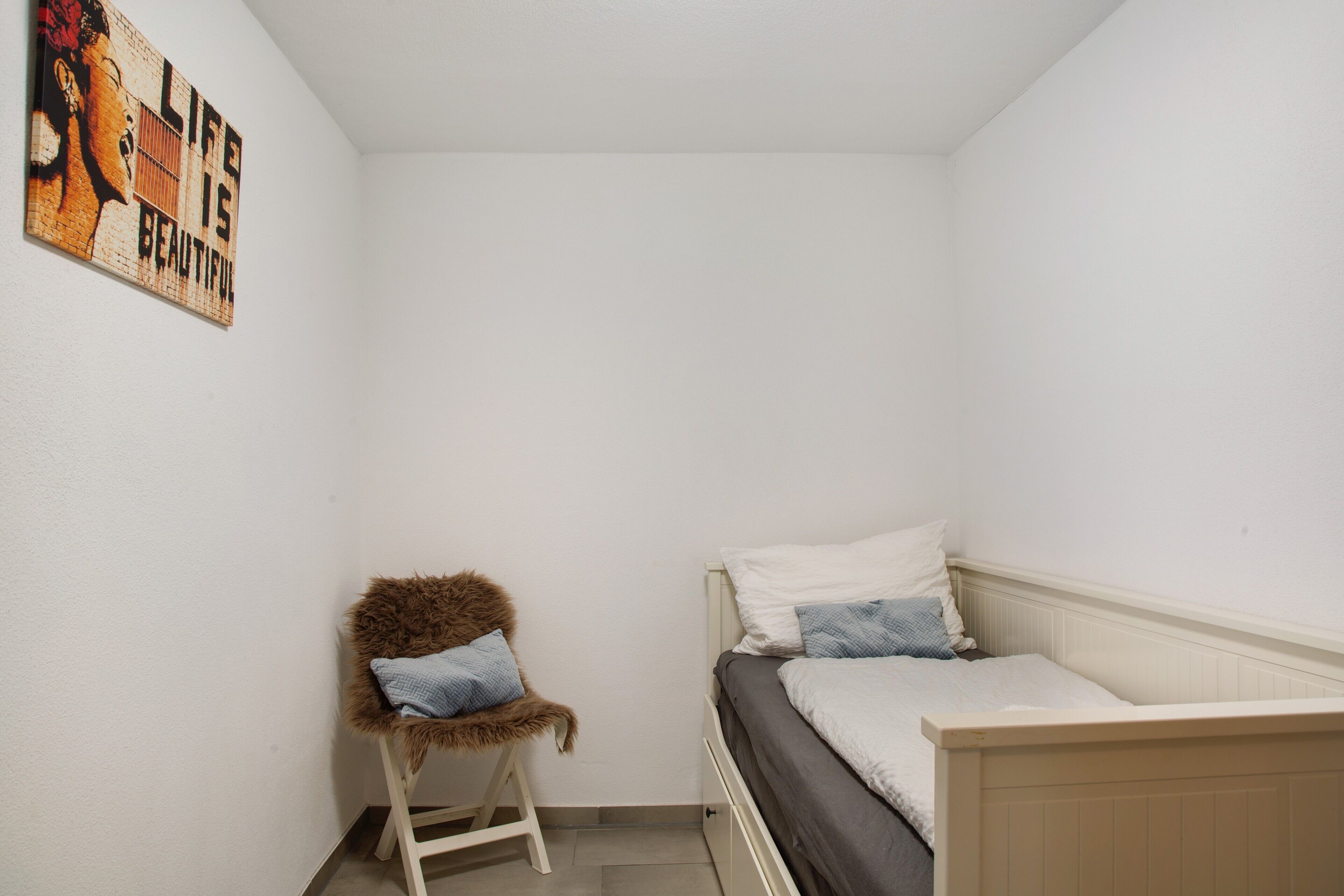 Une petite pièce propre, avec un lit simple, une chaise et une œuvre d’art accrochée au mur.