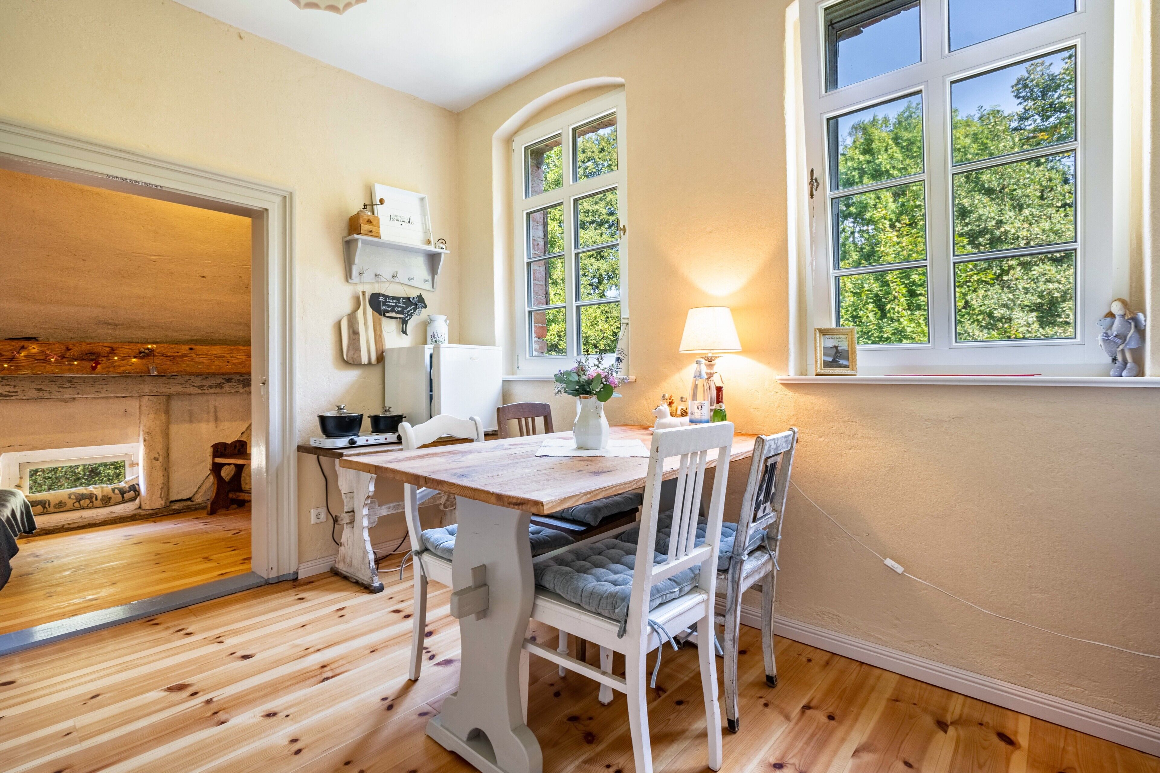 Ein Zimmer mit Holzboden, einem Esstisch mit Stühlen, einem Fenster mit Blick auf Bäume und einer Lampe am Fensterbrett.
