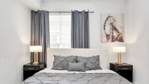 Une chambre à coucher avec un lit, deux tables de chevet avec des lampes, une fenêtre avec des rideaux et un tableau au mur.