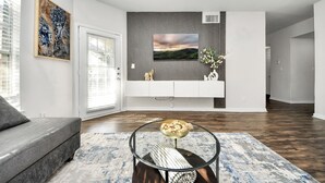 Un salon moderne avec un canapé gris, une table basse en verre agrémentée d’objets décoratifs, un téléviseur fixé au mur et un grand tableau abstrait.