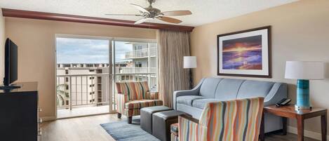 A hotel room with a balcony, a sofa, striped armchairs, a TV, and a painting of a sunset.