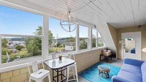 A sunlit room with a table and chairs, a blue sofa, a small stool, and a view of a residential area through large windows. - #StayinMyDistrict Historic Heritage Walk to Bay (Coos Bay)