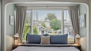 A bedroom with a bed, two bedside tables with lamps, a window with curtains, and a view of buildings.