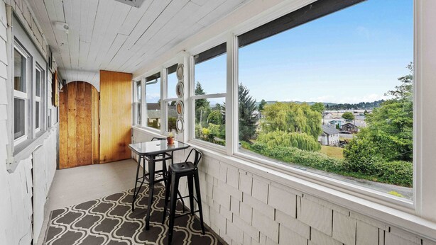 A balcony with a table and chairs, large windows with a view of greenery and buildings, and a wooden door.