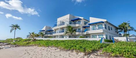 A multi-story beachfront hotel with balconies, palm trees, and a sandy beach in the foreground.