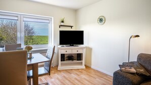 Ein Wohnzimmer mit Fernseher, einem Esstisch, einer Couch und einem Fenster mit Blick auf Bäume.