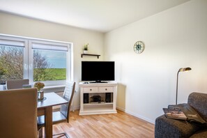 Un salon comprenant un téléviseur, une table à manger, un canapé et une fenêtre donnant sur des arbres.