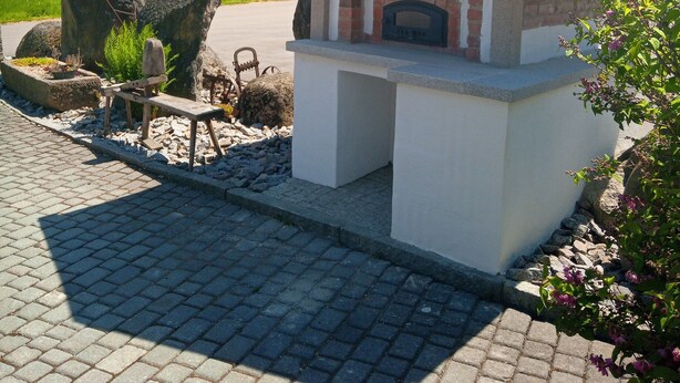 A small outdoor brick oven structure with a chimney, surrounded by a paved area and greenery.