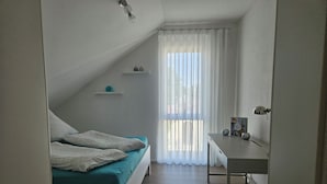 A small, well-lit room with a bed, a desk, and a window with sheer curtains.
