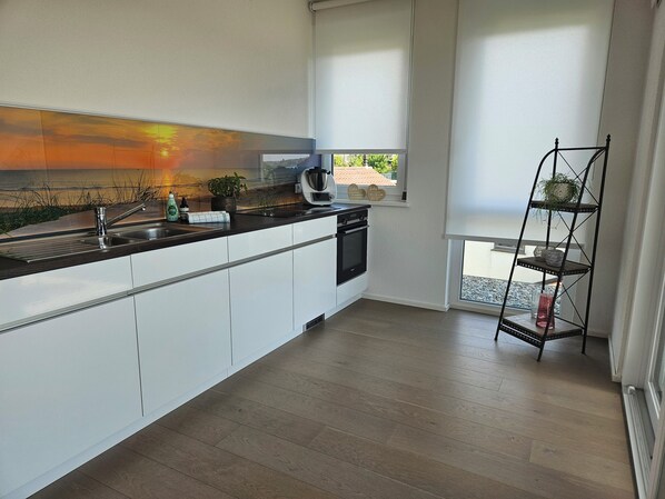 A modern kitchen with white cabinets, a black countertop, and a built-in oven. There is a window with a view of a garden and a shelf with plants. - Sehr Attraktive Luxuriöse Wohnung mit Herrlichem Alpenblick! (Markdorf)