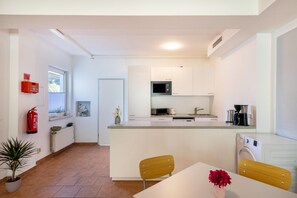 A compact kitchen with a microwave, sink, and washing machine, a dining area with chairs, and a potted plant.