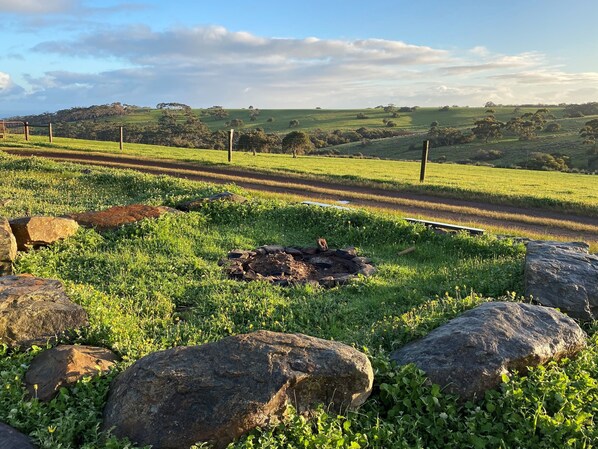 A rural landscape with a grassy field, a dirt road, and a fire pit surrounded by rocks.