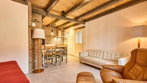 Ein gemütliches Zimmer mit einer Couch, einem Sessel, einem Esstisch mit Stühlen, einer Küchenzeile und einer Holzbalkendecke.