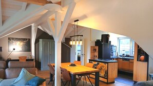 Een open leef- en eetruimte met een houten eettafel, stoelen en een bank met kussens. Achtergrond is een keuken met een koelkast en een raam.
