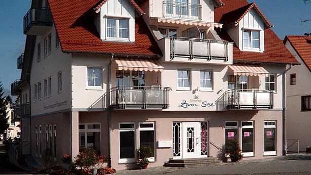 Un immeuble à plusieurs étages, doté d’un toit recouvert de tuiles rouges, de balcons et d’une enseigne sur laquelle on peut lire « Zum See ».
