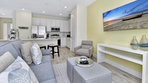 Ein modernes Wohnzimmer mit einem grauen Sofa, einem gestreiften Hocker, einem weißen Couchtisch und einer Küche im Hintergrund.