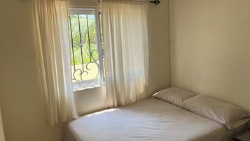 Un lit simple avec un matelas et un oreiller blancs, une fenêtre avec des rideaux blancs et un appareil de climatisation fixé au plafond.
