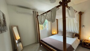 A bedroom with a four-poster bed, a bedside table with a lamp, a mirror, and a window with curtains.