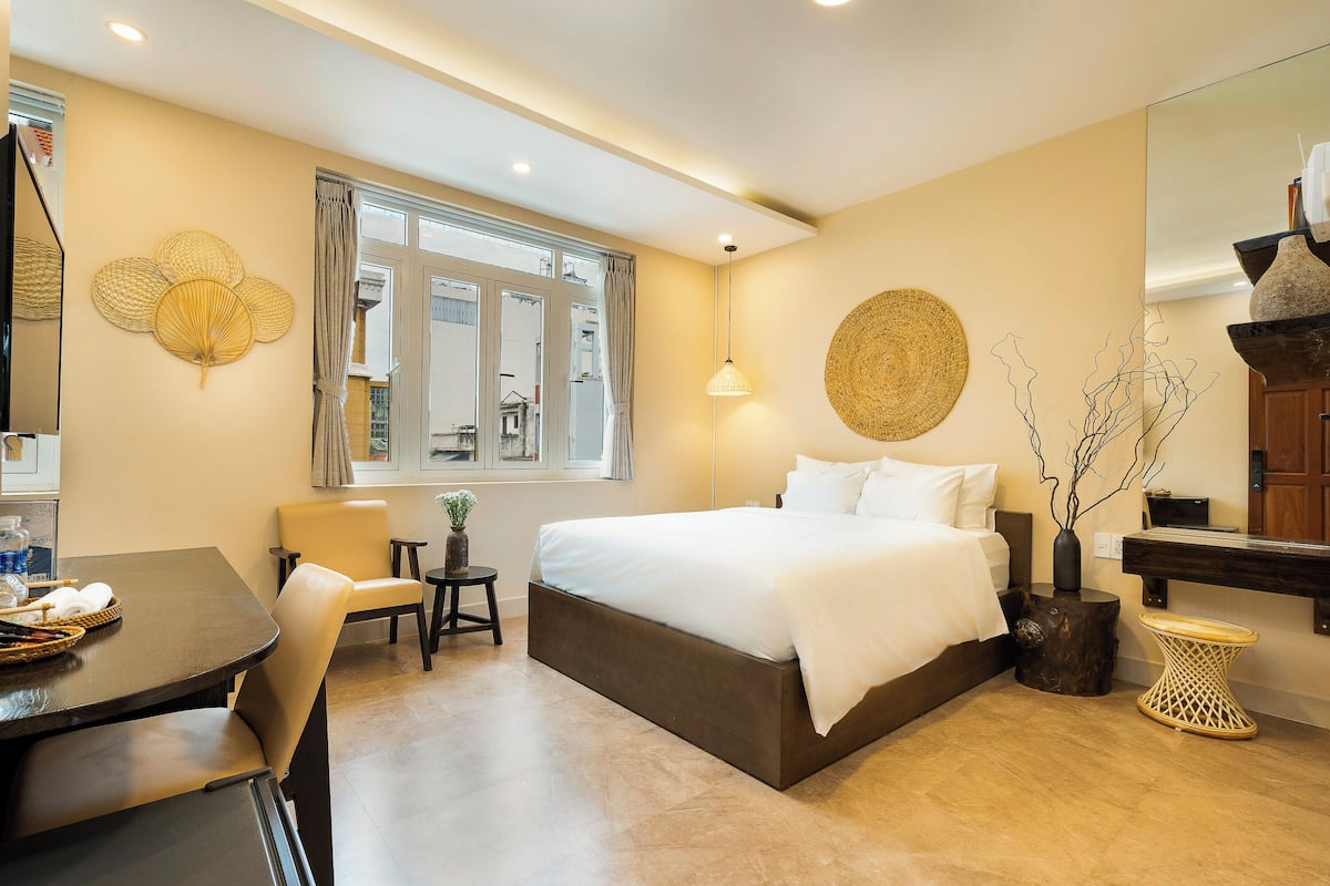 A hotel room with a large bed, a dining table, a chair, a small side table, and a large window.