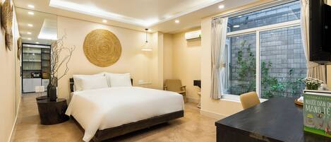A modern bedroom with a large bed, a desk, and a window with a view of greenery.