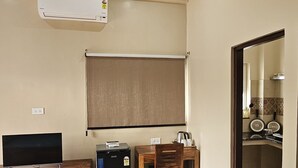 Una habitación con televisión de pantalla plana, un refrigerador pequeño, una mesa de madera con una silla y un ventanal con persiana.