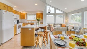 A modern kitchen with wooden cabinets, a central island, and a dining area with a table set for breakfast. - Ocean Views, Game Garage & Steps to Neahkahnie Beach | Bella Sol by AvantStay (Nehalem)