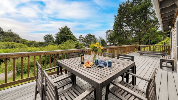 A wooden deck with a table set for dining, surrounded by chairs, and overlooking a lush landscape. - Ocean Views, Game Garage & Steps to Neahkahnie Beach | Bella Sol by AvantStay (Nehalem)