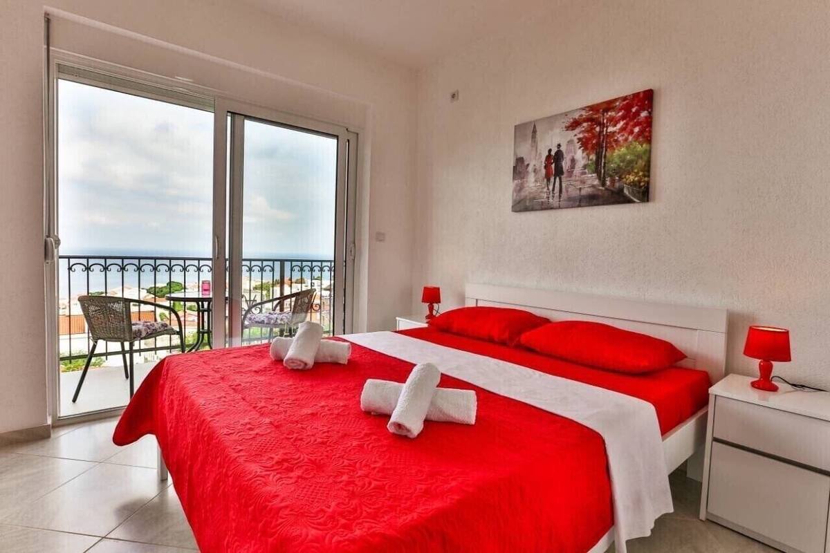 A hotel room with a large bed, red bedding, a balcony with a view, and a painting on the wall.