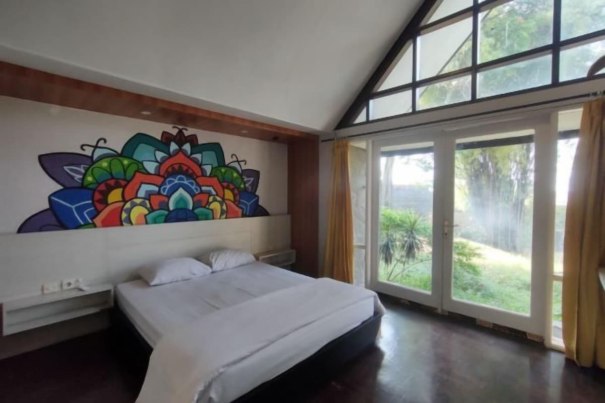 A bedroom with a large bed, a colorful mural on the wall, and a sliding glass door leading to a garden.
