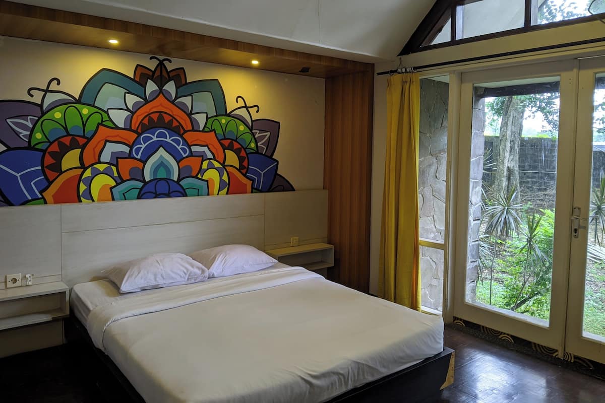 Une chambre avec un lit, une fresque murale colorée, une tête de lit en bois et une vue sur l’extérieur à travers une porte vitrée.