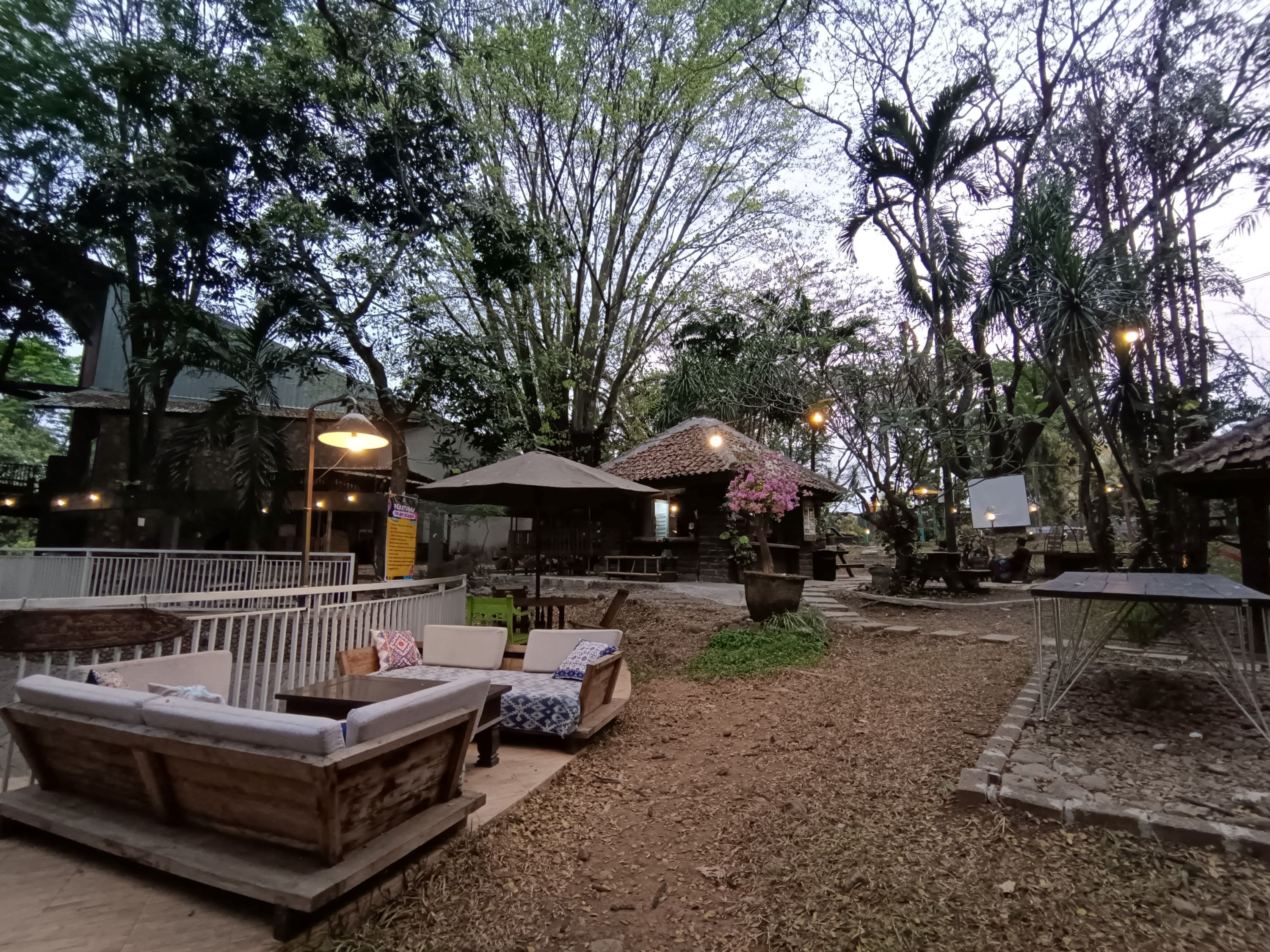 An outdoor seating area with wooden benches and tables, surrounded by trees and a small building with a tiled roof.