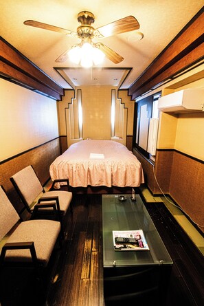 A compact hotel room with a bed, two chairs, a glass table with a magazine, a ceiling fan, and a window. - Fullsfar Hotel (Kyoto)