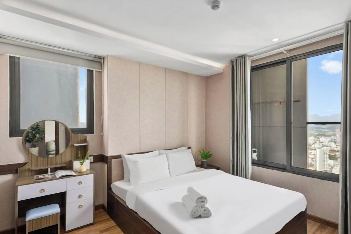 A hotel room with a bed, a vanity with a mirror, a small desk, and a window with a city view.