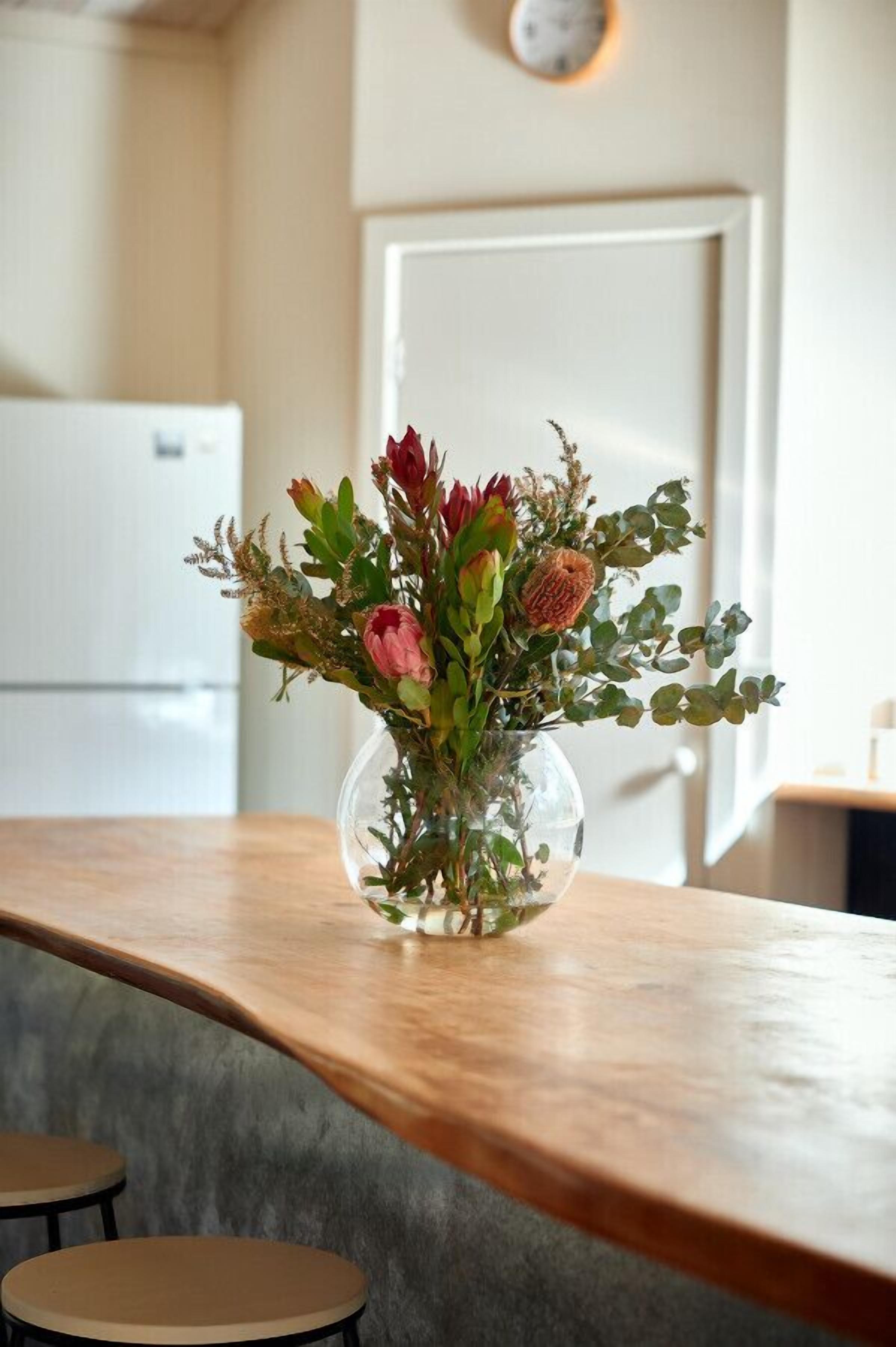 Eine klare Glasvase mit Blumen auf einem Holztisch in einem Raum mit einer weißen Tür und einer Uhr an der Wand.