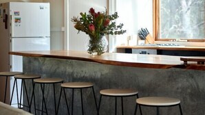 A kitchen with a wooden counter, bar stools, a refrigerator, a clock, and a window with a view of trees. - Rustic Treehouse - Aireys Inlet (Aireys Inlet)
