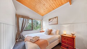 A bedroom with a large bed, wooden ceiling, white walls, a window with curtains, a bedside table with a lamp, and a painting on the wall. - Rustic Treehouse - Aireys Inlet (Aireys Inlet)