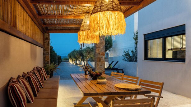 An outdoor dining area with a wooden table and chairs, a cushioned bench, and hanging lamps. - GEMM Beach House - Beachfront Apartment in Hersonissos (Hersonissos)
