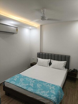 A hotel room with a bed, a ceiling fan, an air conditioning unit, a bedside table with a phone, and a nightstand. - Hotel Sunrise Ecostay  (New Delhi)