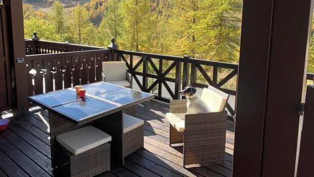 A deck with wicker furniture and a glass-top table, overlooking a mountainous landscape with autumn foliage.