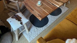 A cozy living room with a wooden coffee table, two chairs with fur cushions, a sofa, and a kitchen area with a shelf above the counter.