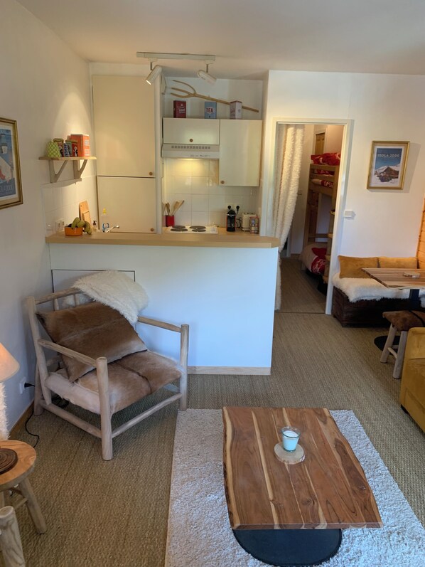 A cozy living room with a wooden coffee table, a plush chair, and a small kitchen area with a fridge and cabinets.