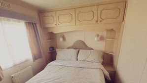 Une chambre à coucher comprenant un lit, un radiateur, une fenêtre avec des rideaux et une tête de lit dotée d’étagères intégrées.