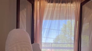 A neatly made bed with white linens and a brown bedspread, a window with sheer curtains, and a view of trees outside. - Sirtaki Apartments (Kassandra)