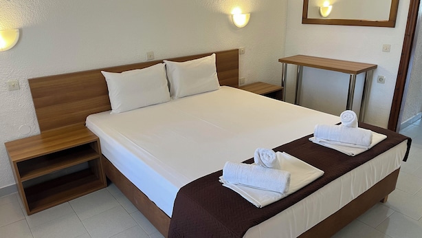 A neatly made bed with white linens and brown bedding, a wooden nightstand, a small bedside table, and a wall-mounted light fixture. - Sirtaki Apartments (Kassandra)