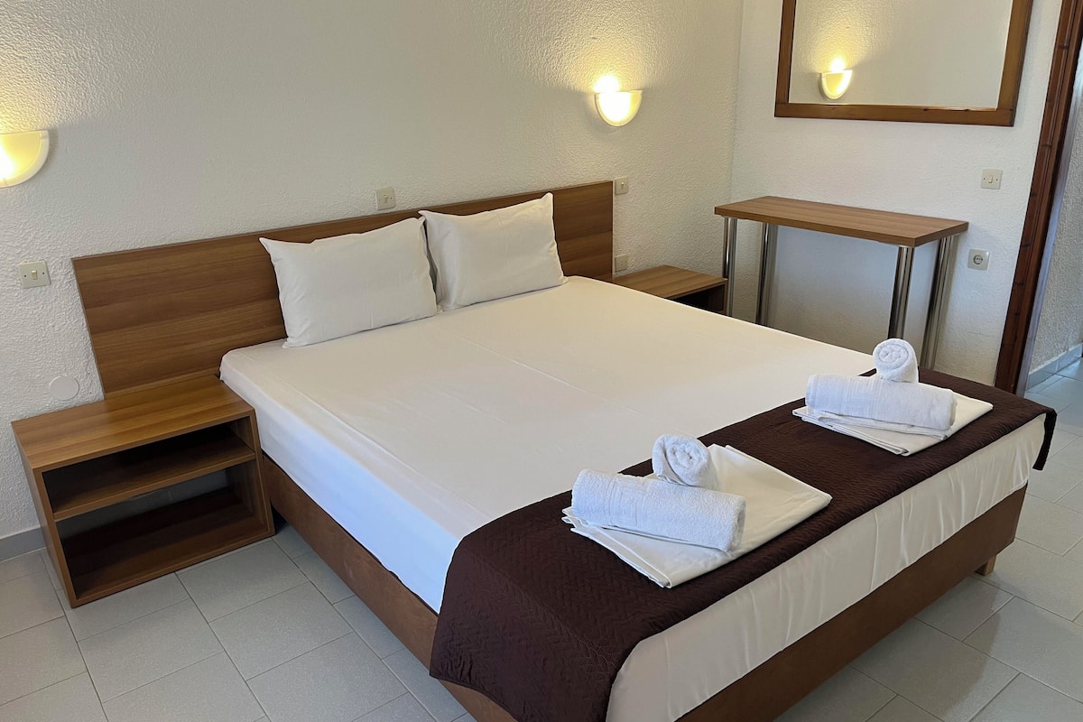 A neatly made bed with white linens and brown bedding, a wooden nightstand, a small bedside table, and a wall-mounted light fixture.