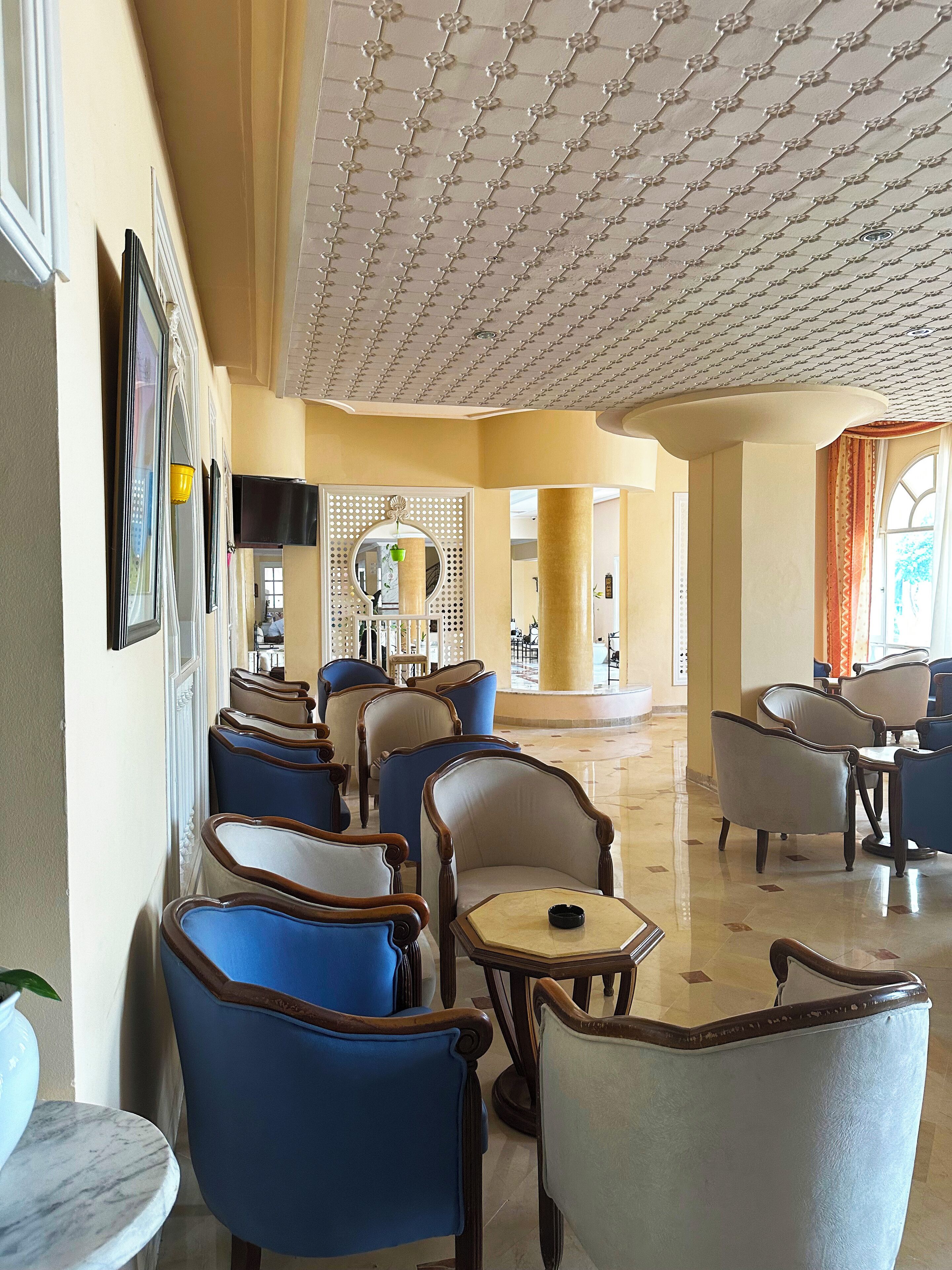Un hall de hotel con filas de sillones azul y beige, una mesa de mármol y un techo decorativo.