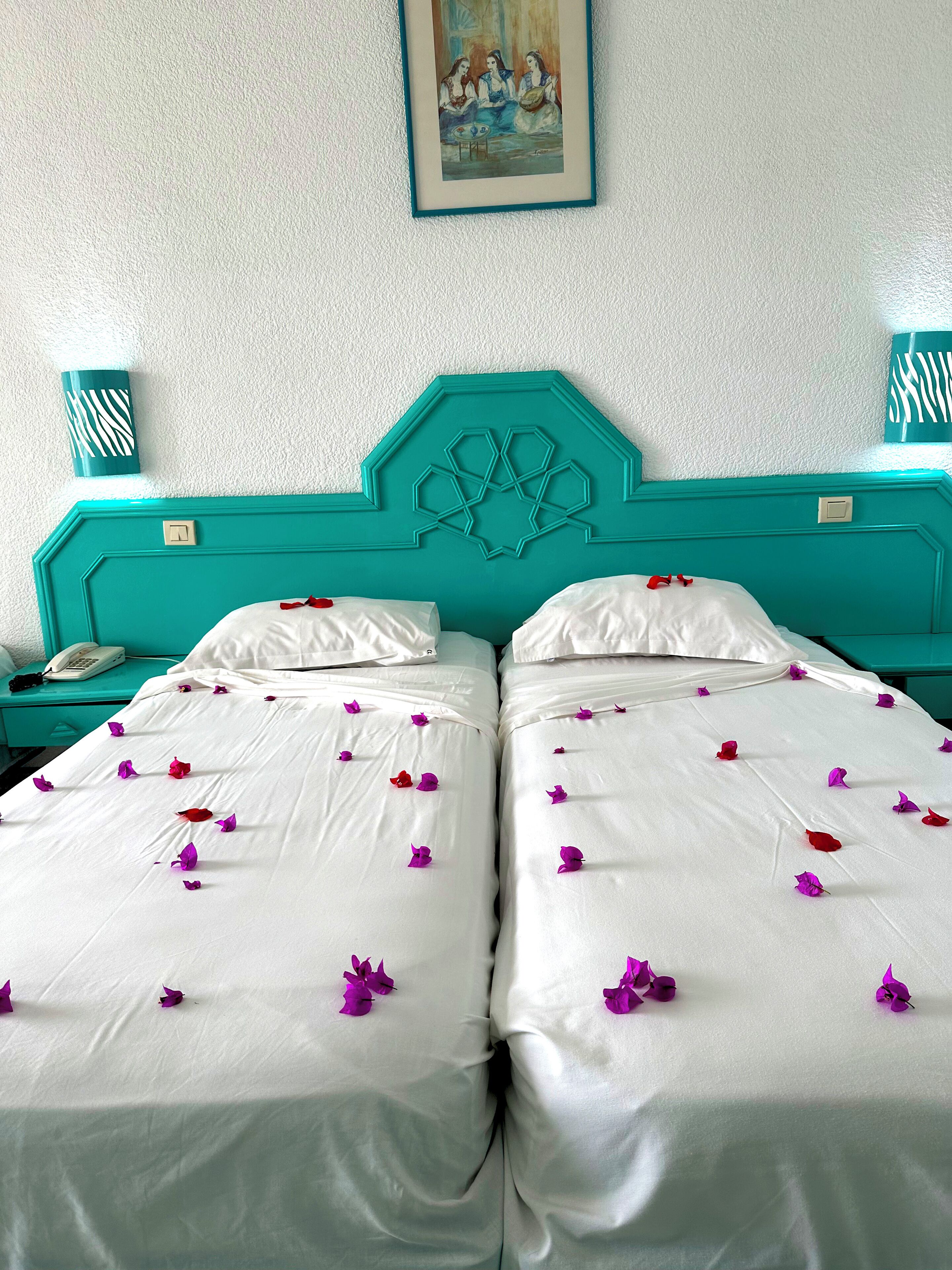 A double bed with white linens and purple flower petals, teal headboard, and a painting on the wall.