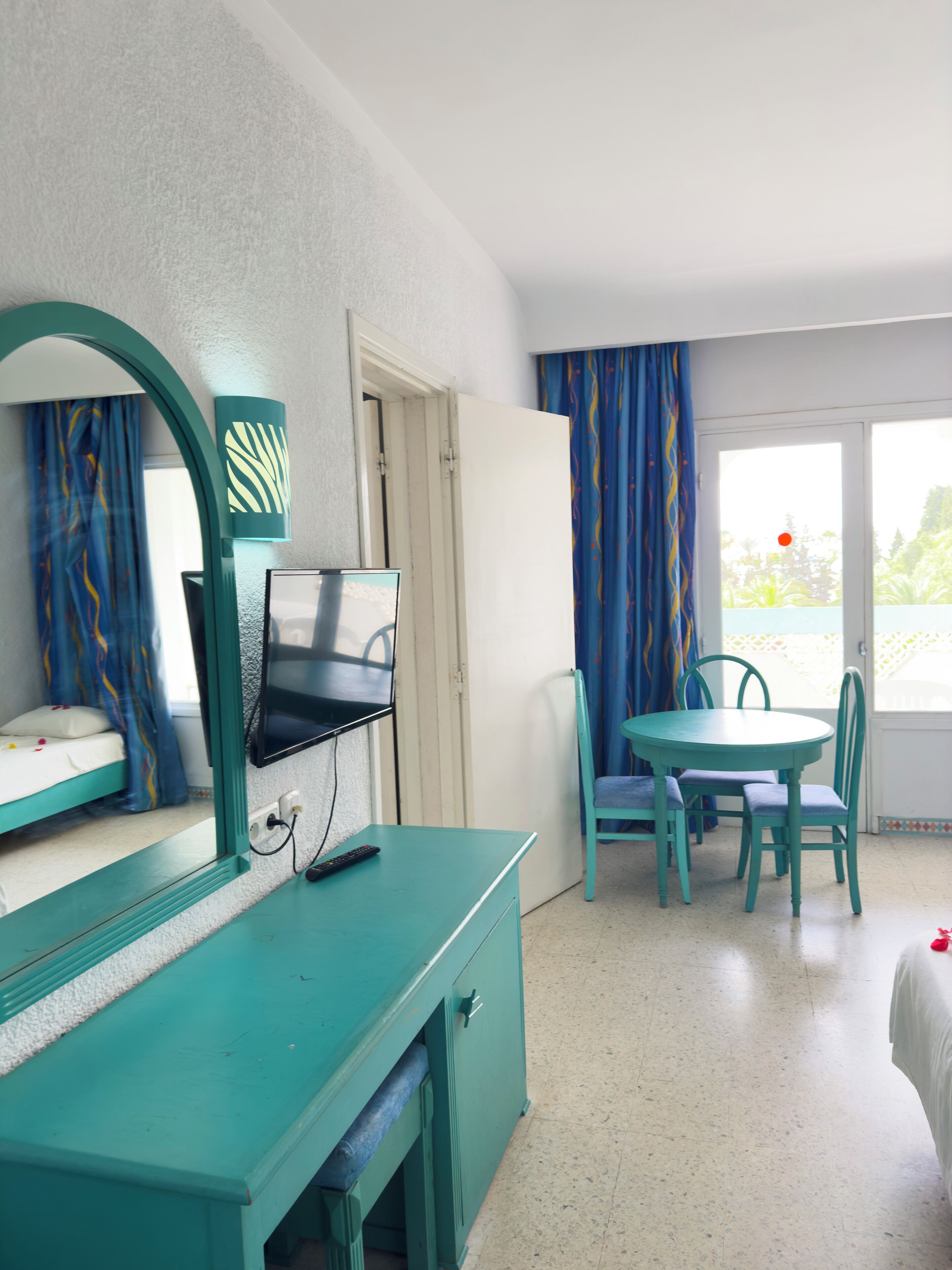Una stanza con un letto a castello, una scrivania con specchio, una televisione, un tavolo da pranzo con sedie e una finestra con tende.