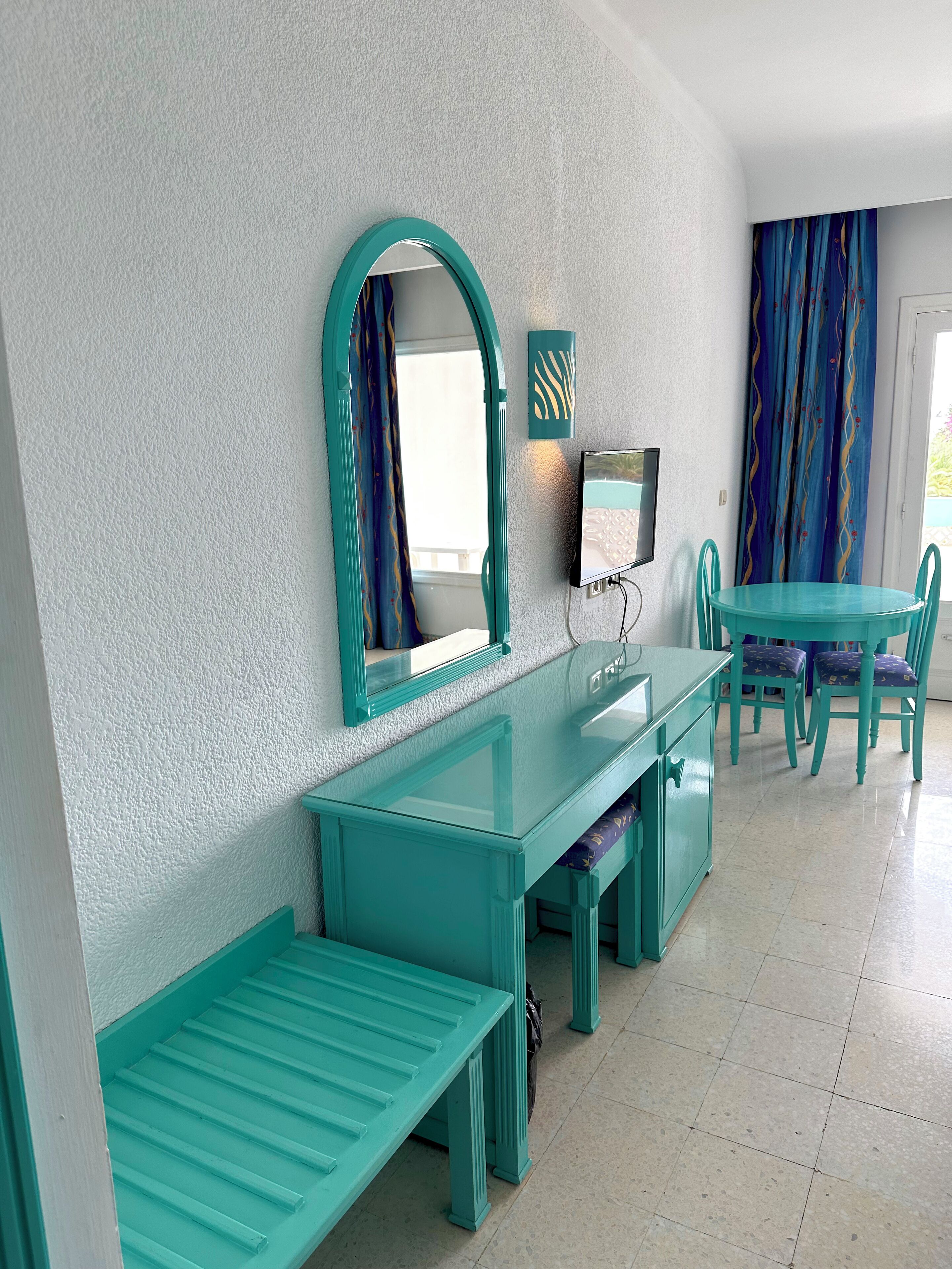 Ein Zimmer mit Türkis-Möbeln, einem großen Spiegel, einem Flachbildfernseher und Blick ins Freie durch ein Fenster.